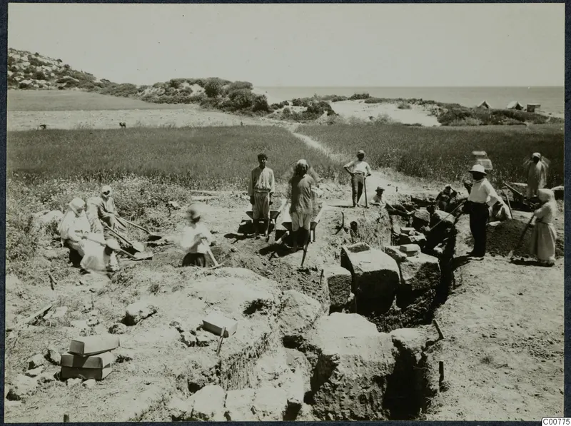 Workers with spades and hoes at an excavated trench among stone blocks, with a green field and the sea behind.