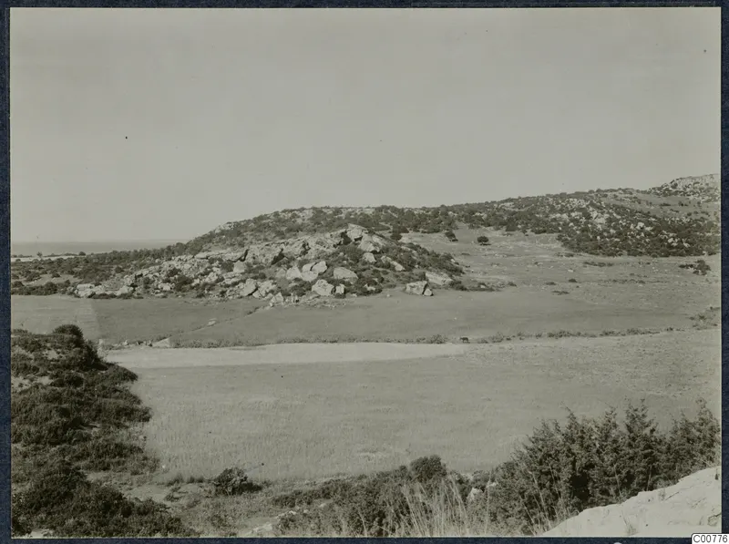 Rocky hills with scrub and open grassy fields near Oura, with the sea visible on the horizon at left.