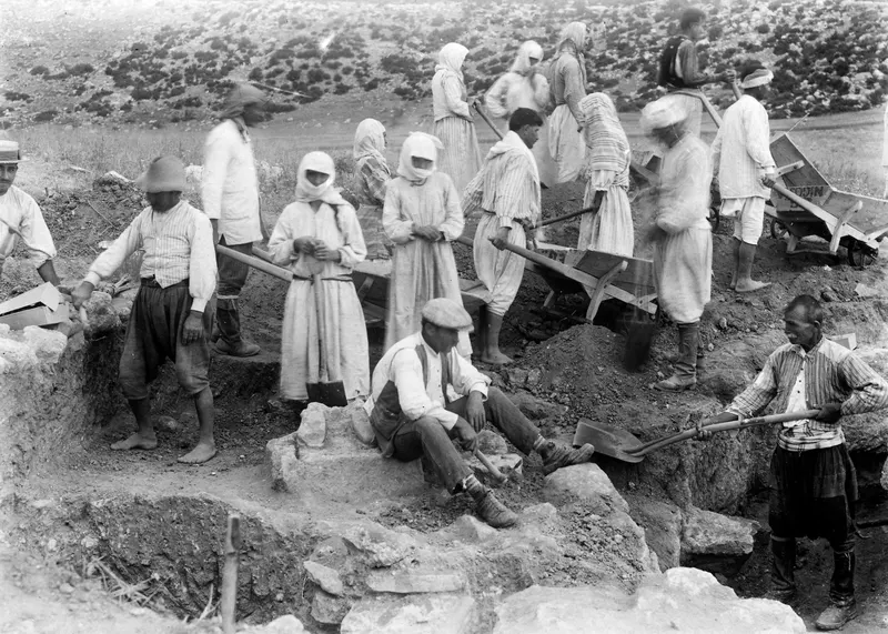 Workers digging an excavation at Oura, with wheelbarrows, shovels and exposed stone walls.