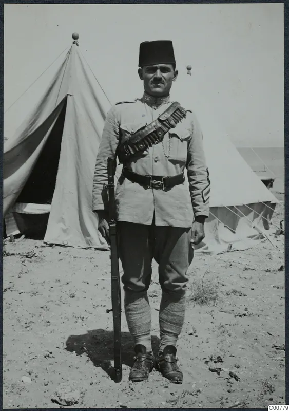 Standing policeman in a fez and khaki uniform holding a rifle, in front of a tent, at Oura