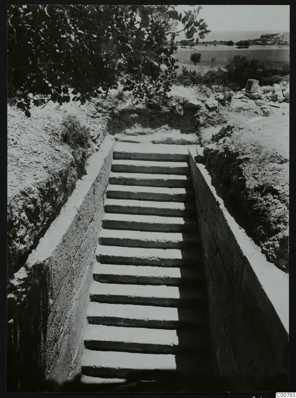 Stepped dromos cut down between walls toward a tomb entrance at Trachonas, with the sea in the distance.