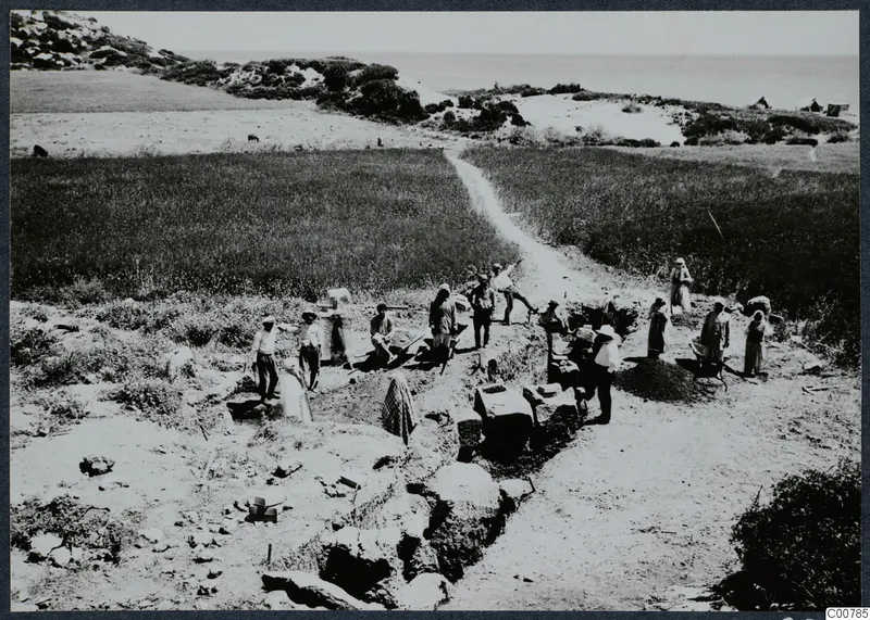 Workers at the Oura excavation, with a trench, exposed stone blocks and wall foundations, beside cultivated fields and the sea.