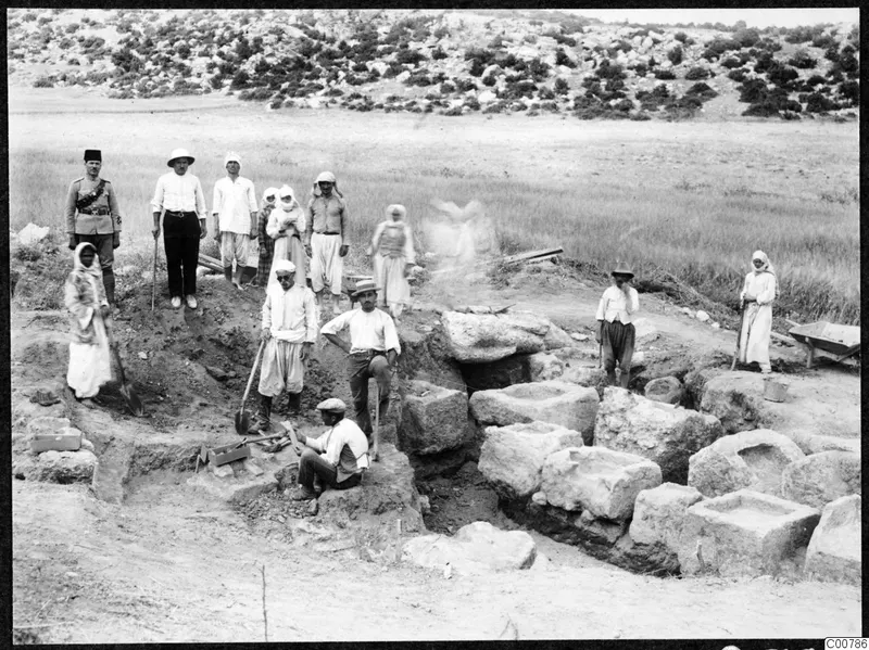 Workers and local people gathered at an excavation among cut stone blocks, with a uniformed man standing at left.