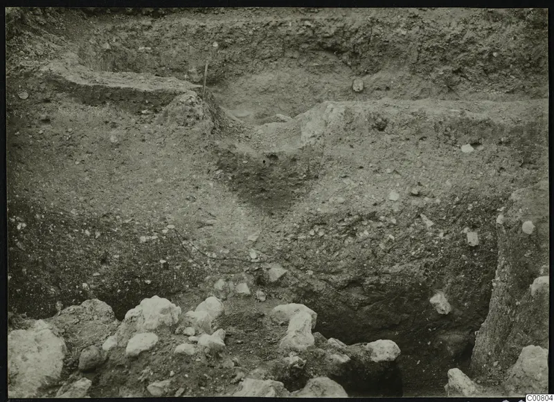 Excavation trench at Plakes, Lapithos, with layered soil faces and pale stones at the bottom