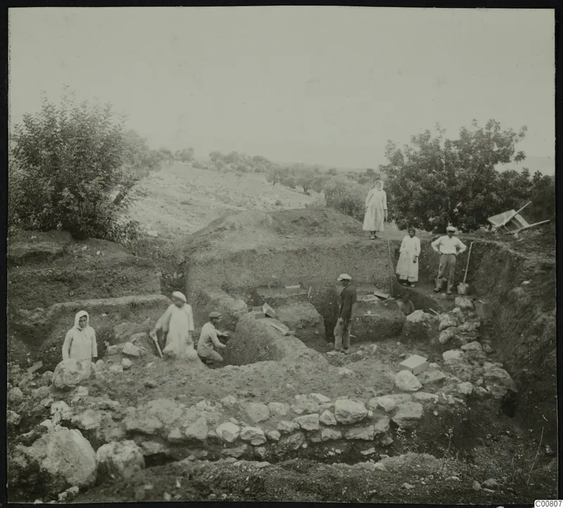 Excavation at Plakes, Lapithos, with workers in a trench beside stone walls, and a wheelbarrow on the bank at right.