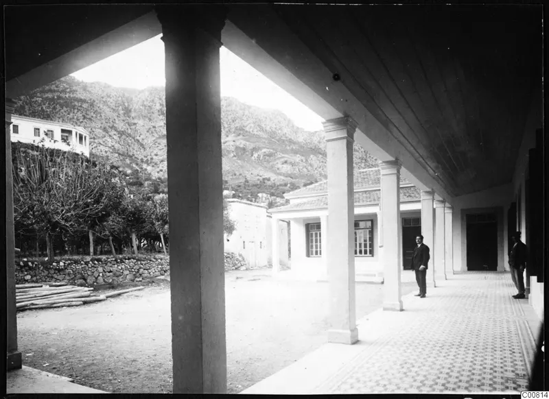 Two men standing in a colonnade, with a building, a stone wall and a mountain behind