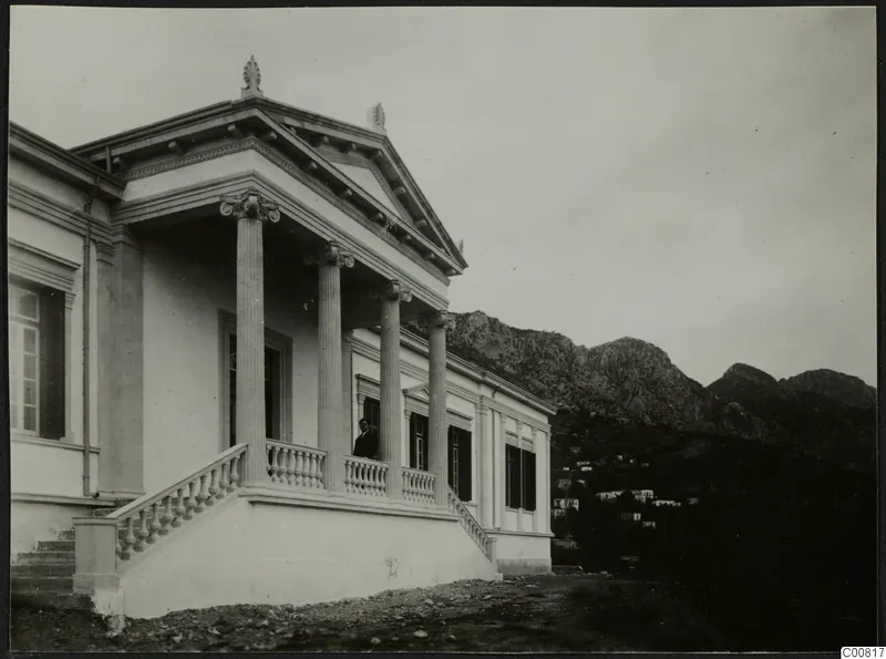 Building with a columned porch and pediment, a man standing on the terrace, mountains rising behind