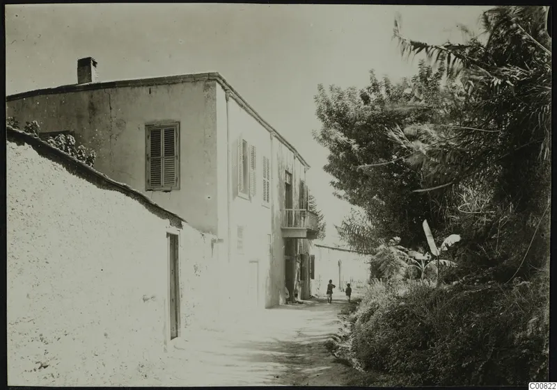 Whitewashed two-storey building beside a dirt lane at Lapithos, with trees on the right and two people on the track