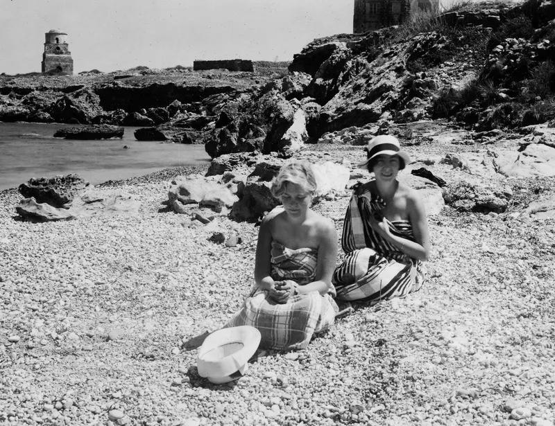 Two young women wrapped in towels sit on a pebble beach beside rocks and the sea at Lambousa.
