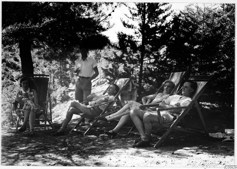 Five people resting in deckchairs under pine trees outside the expedition house at Machairas.
