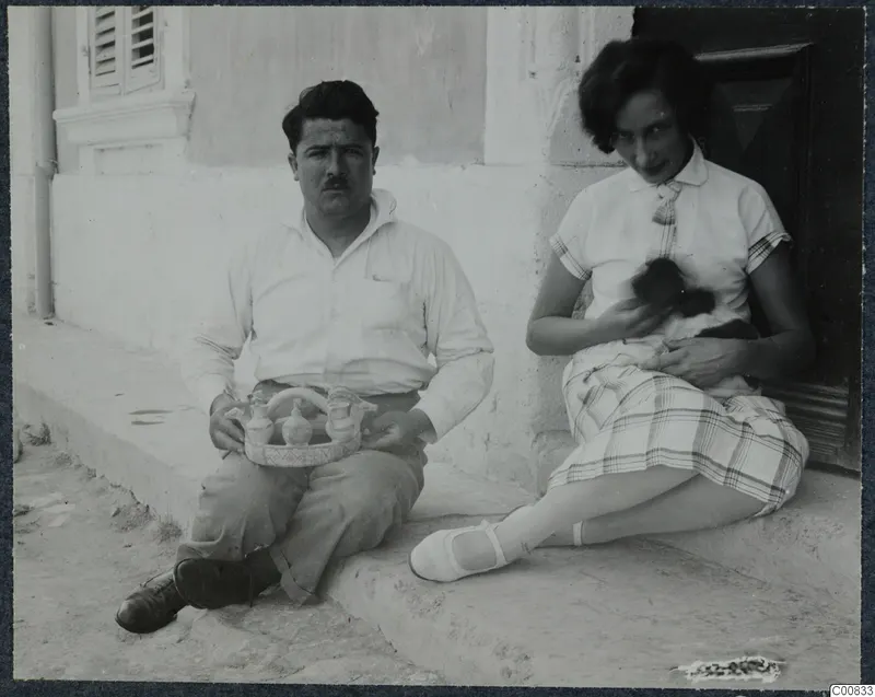 A man and a woman sitting on a whitewashed doorstep; he holds a tray of small vessels and she holds a cat.