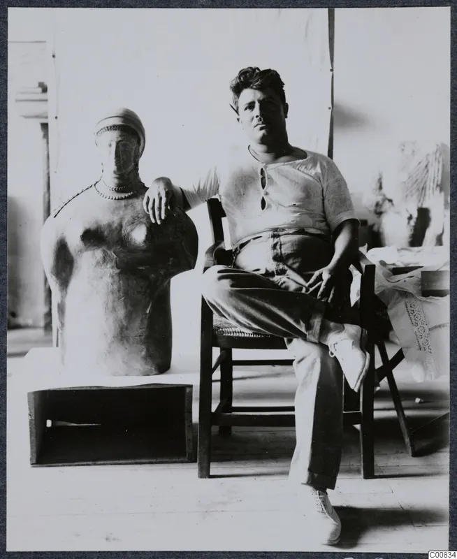 Toulis Souidos seated beside a large sculpture of a figure with a beaded necklace and cap