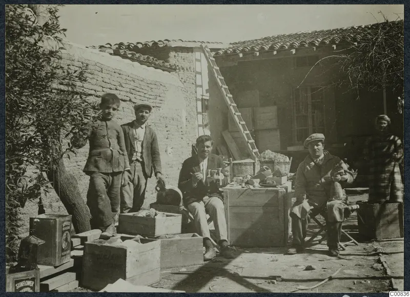 Courtyard with five people among wooden crates, jugs and vessels, a ladder leaning on a wall and a tiled-roof building behind
