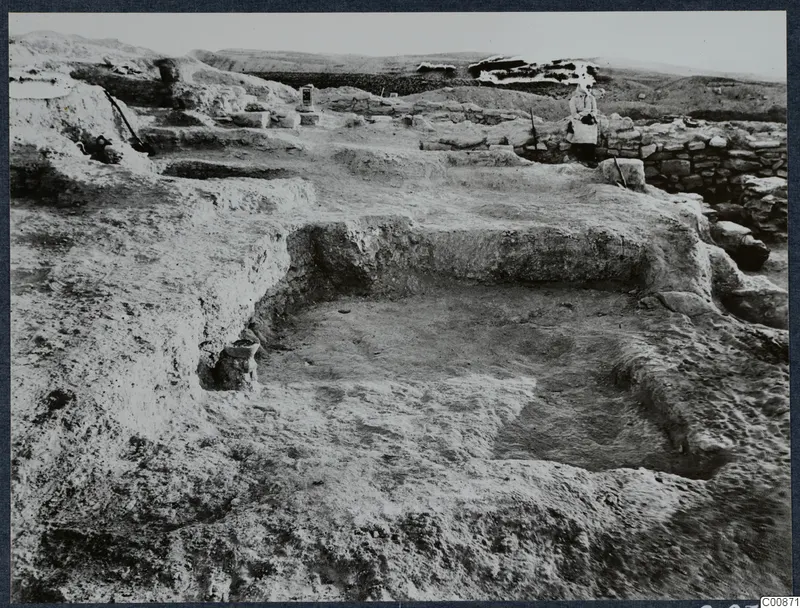Excavated square pits at Idalion with a low stone wall and a seated person beyond, seen across the dig