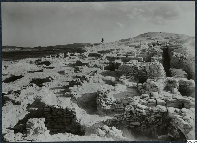Excavated stone walls and foundations at Idalion Acropolis, with two people on the ridge and bare hills behind.