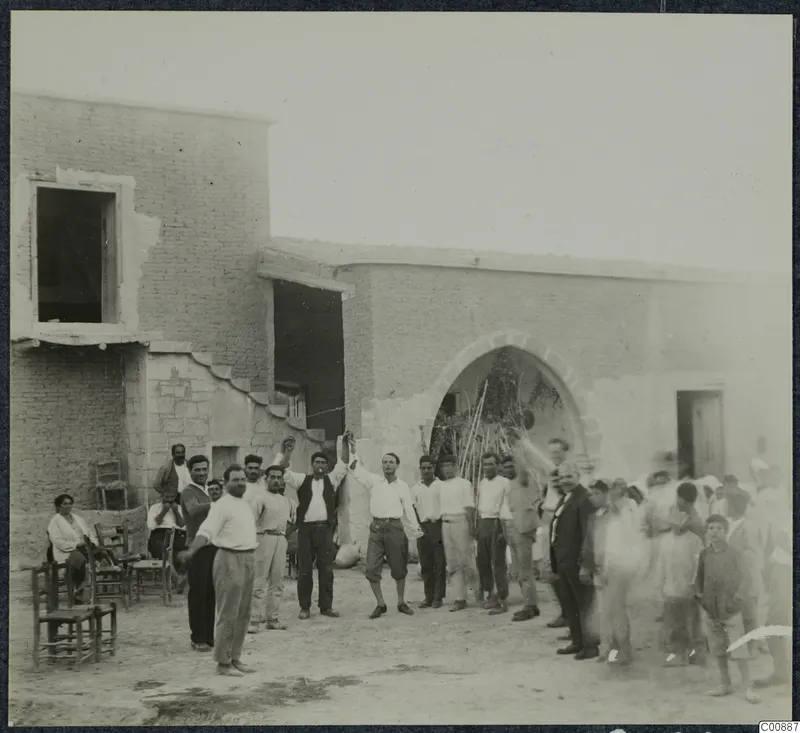Farewell party at Panajiotis, Idalion: a crowd of men gathers in a dusty yard before a mud-brick building with an arched doorway