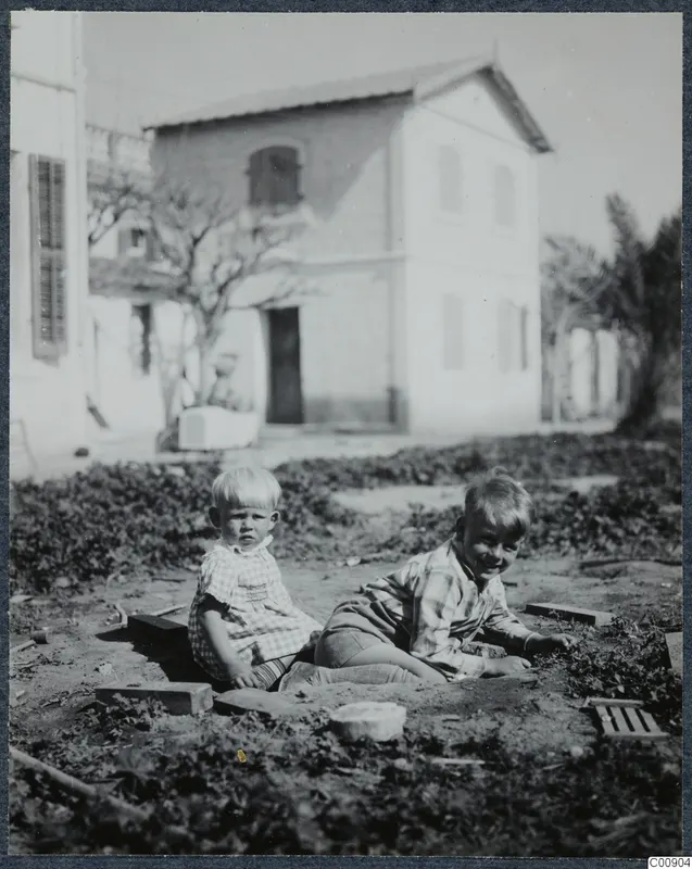 Two young children sitting on bare ground in front of a whitewashed two-storey building, Nicosia