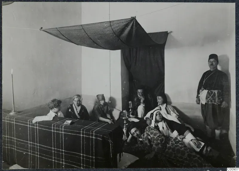 Staged "Oriental" party scene: people in dress-up clothes around a checked-covered bed under a draped dark cloth.