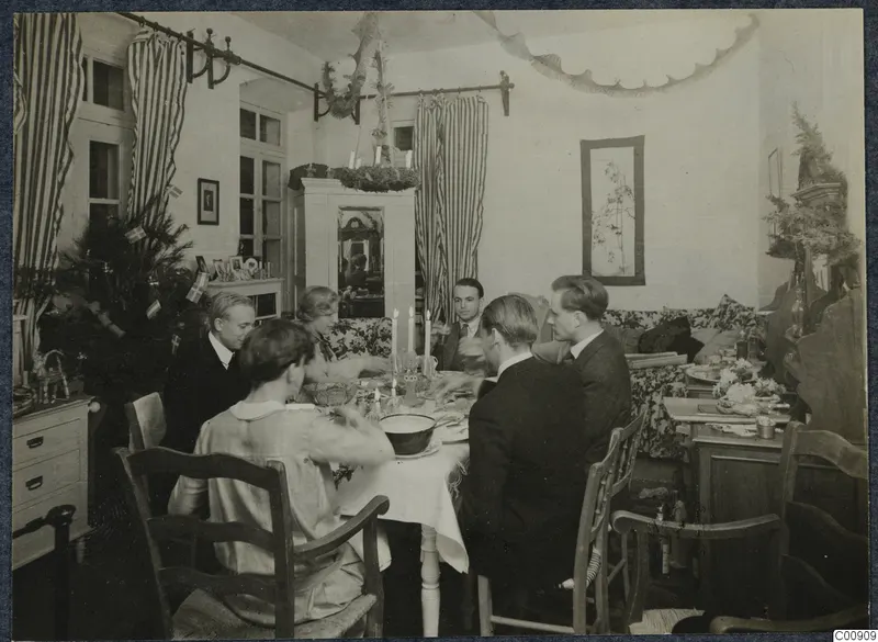 Christmas Eve 1927 meal: people seated at a candlelit table in a decorated room with striped curtains and a Christmas tree with flags