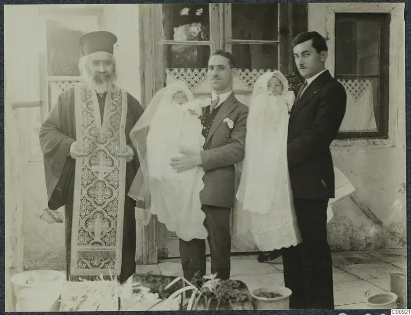Priest and two men holding babies in white christening robes in front of a glazed wooden door, with bowls and greenery in the foreground