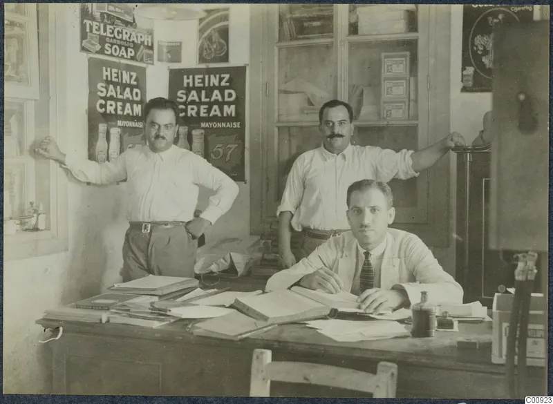Office interior at Kalavasos with a seated man writing at a desk, two men behind him and a Heinz Salad Cream advertisement