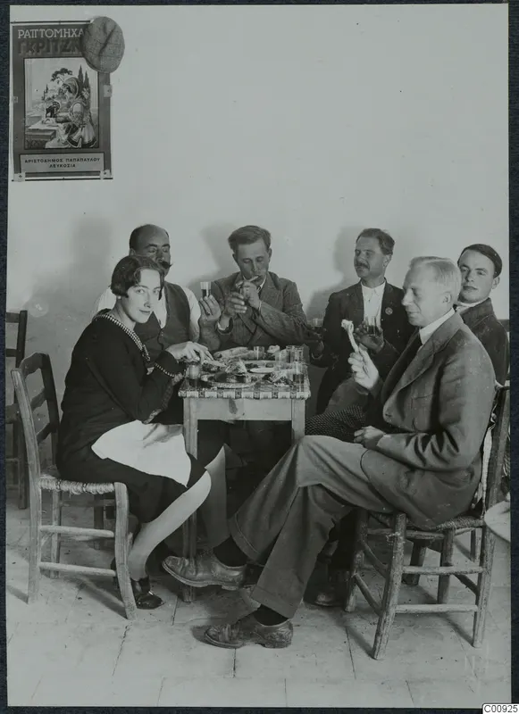 Six people eating and drinking around a small table in a whitewashed room in Lapithos