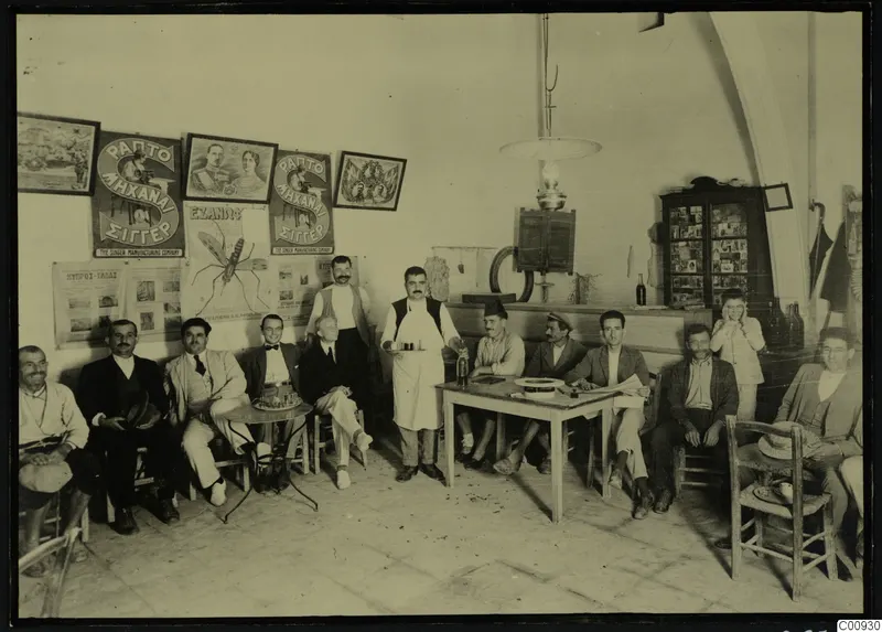 Group of about a dozen men and a waiter in a coffee house interior at Lapithos, seated and standing around tables.