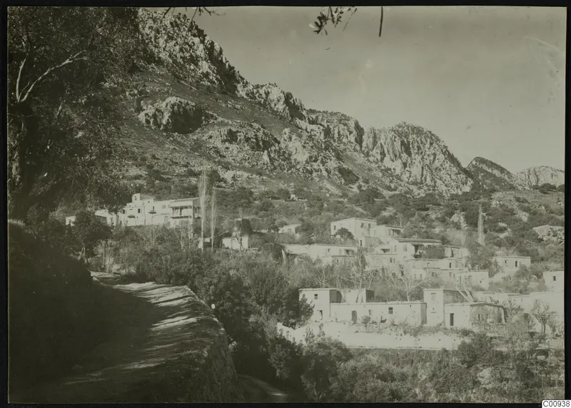 Village of white houses on a hillside below rocky mountains, with trees in the foreground and a track along a retaining wall.