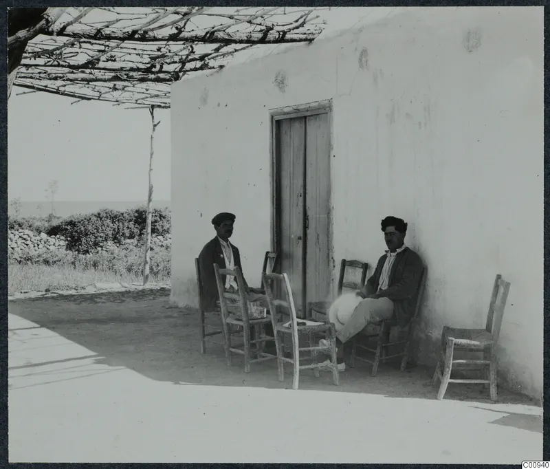 Two men sit on wooden chairs outside a whitewashed building at Karavas, beside a wooden door and a vine pergola.