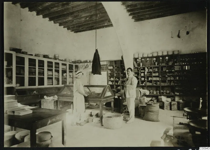 Rosa Lindros and Perikles Kaplanis standing in a storeroom at Lapithos with glass cabinets and shelves of pottery
