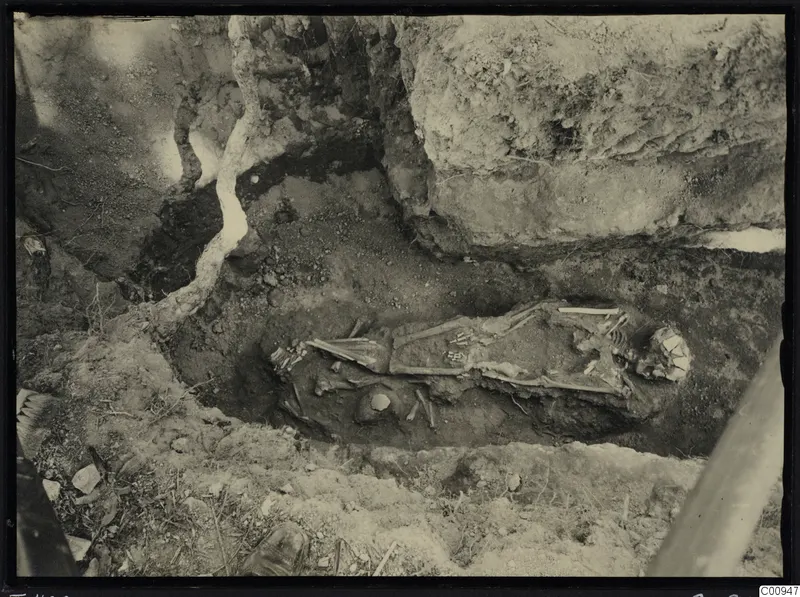 Skeleton lying in an excavated trench at Lapithos, with bones exposed in dark soil beside a large rock face.