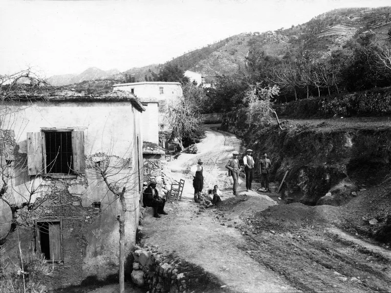 Village street at Kylistra, Lapithos, with whitewashed houses, several people and an excavated earth bank at right
