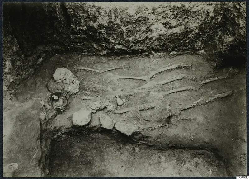 Skeleton lying in an excavated pit at Idalion, with small vessels beside it and a rock face above