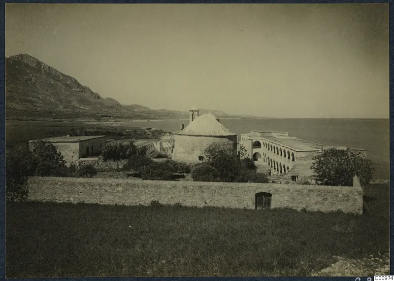 Monastery buildings at Lambousa seen from a walled field, with a domed round structure and arcaded range beside the sea.