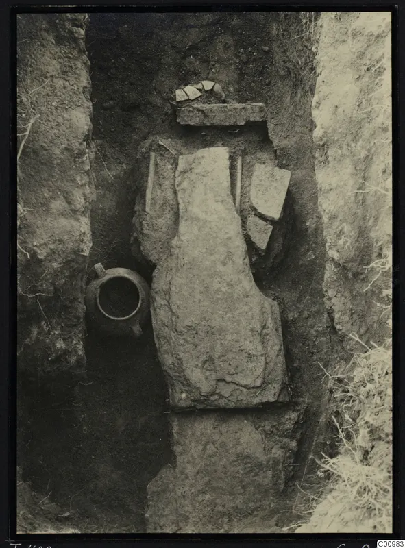 Tomb 422 at Lapithos Kastros during excavation, with a large stone slab, a handled jug and sherds in the cut.