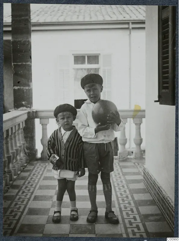 Two young boys standing on a tiled veranda, one holding a large ball, in Nicosia