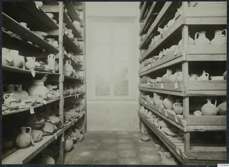 Storage room with wooden shelves on both sides filled with pottery jugs and bowls, many with paper tags, and a window at the back.
