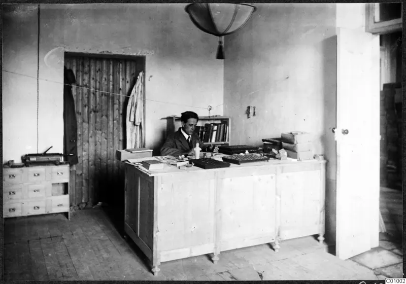 Einar Gjerstad seated at a wooden desk in an office room in Nicosia, with a door and coat behind him.