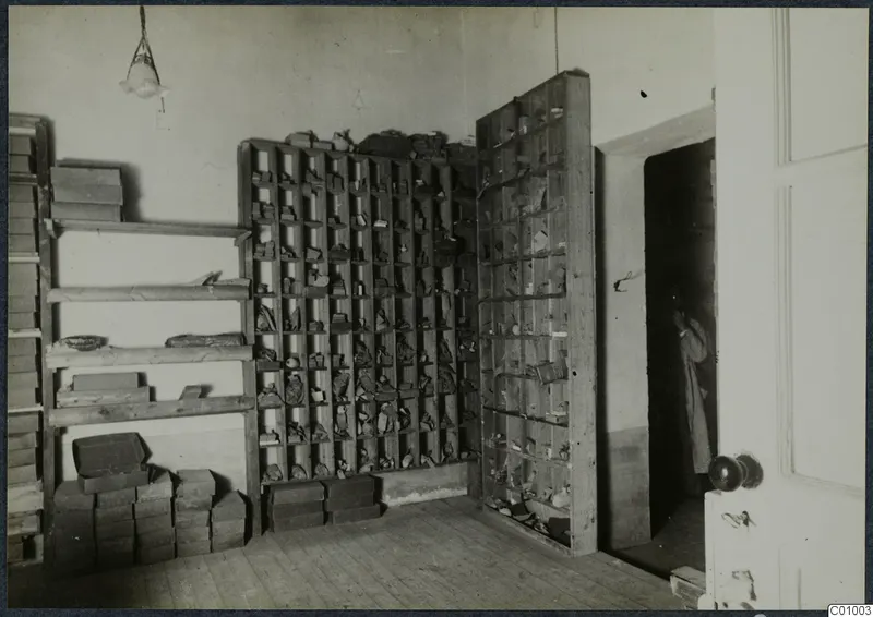 Room with tall wooden compartmented racks holding small objects and stacked boxes; a person stands in the doorway.