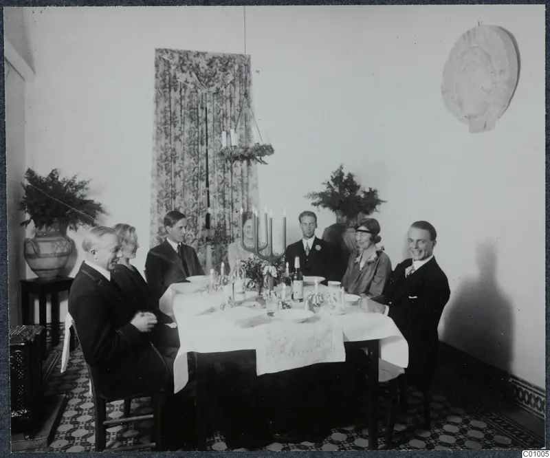 Christmas Eve 1928 dinner: seven people seated at a table with a candelabra, bottles and a white cloth.