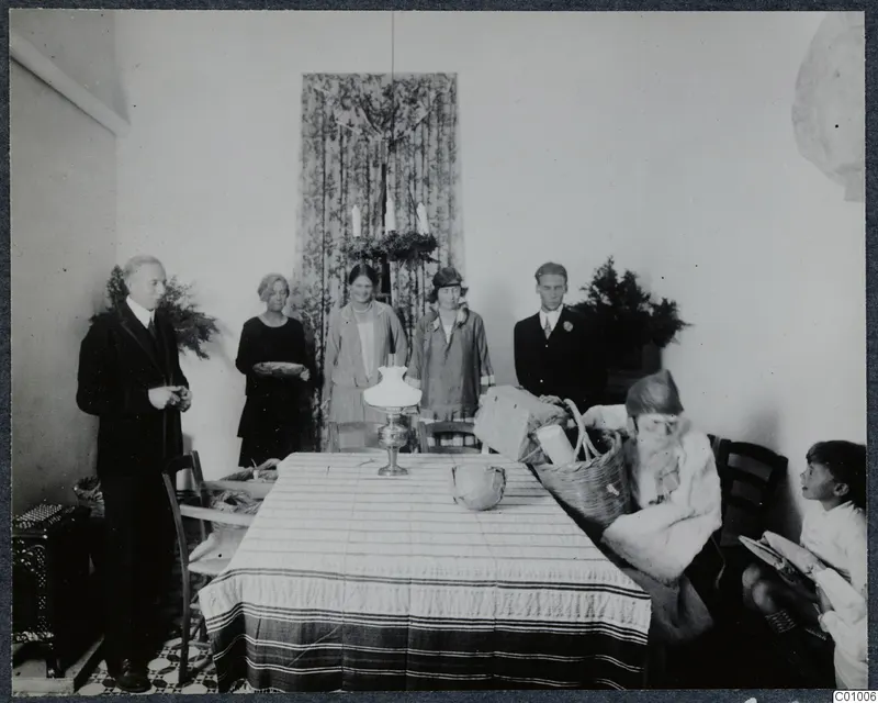 Christmas Eve 1928: adults stand in a room while a figure in fur sits at a table with a basket and parcels; a child sits at right.