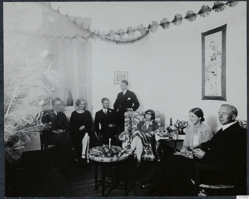 Seven people in a decorated living room on Christmas Eve, with a fruit basket and a lit candle on a low table.
