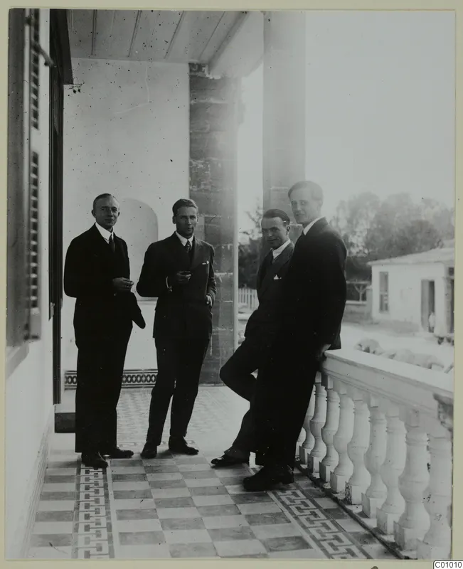 Four men in dark suits standing on a tiled veranda beside a stone column and white balustrade, Nicosia.
