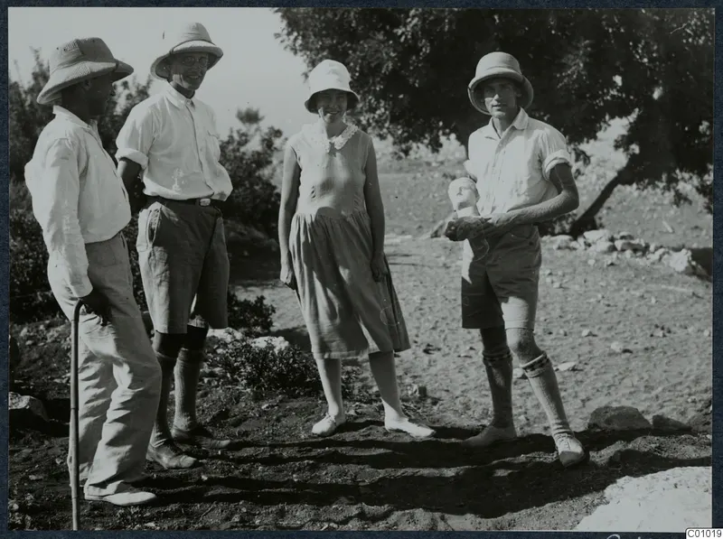 Four people in pith helmets and light clothing standing on dry ground at Vouni, one holding a small pale object.