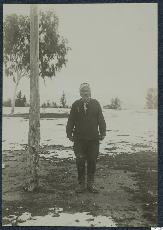 Man in a striped turban and dark jacket standing on patchy snow beside a tall bare tree trunk, Nicosia
