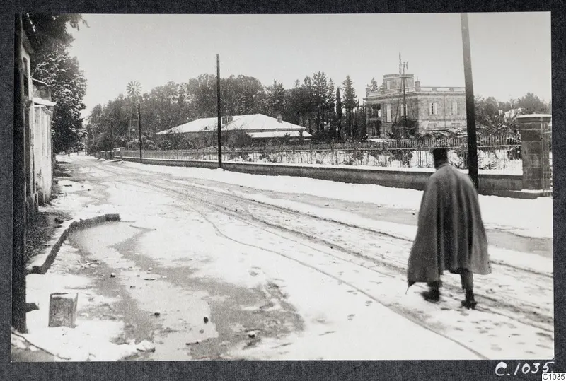 Snow-covered Gladstone Road in Nicosia, with a man in a long cape walking away along the street