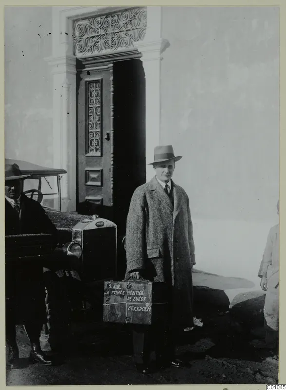 Einar Gjerstad in a tweed overcoat and hat, carrying a box, beside a car and a carved wooden doorway in Nicosia.