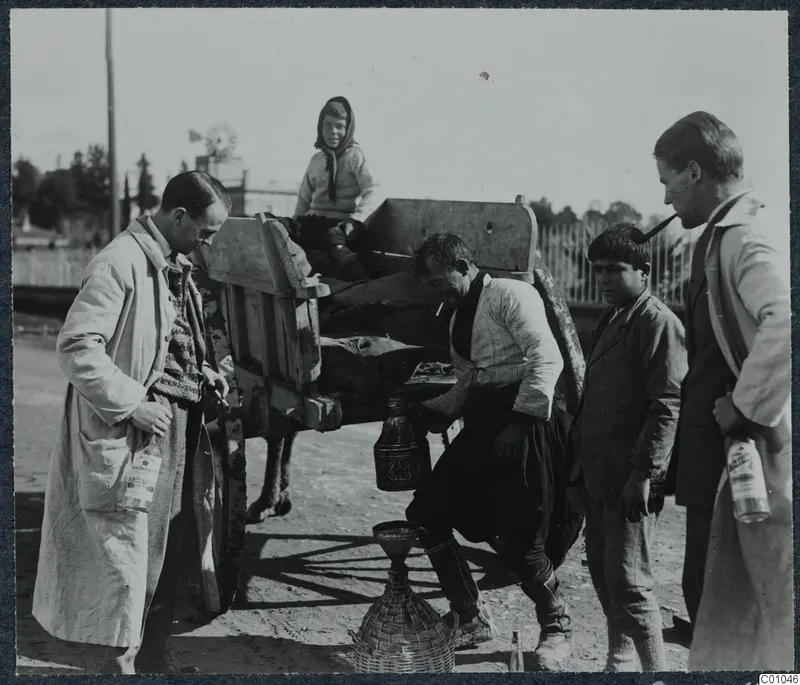 Men and children at a cart in a street, one man crouching over a wine demijohn, Nicosia.