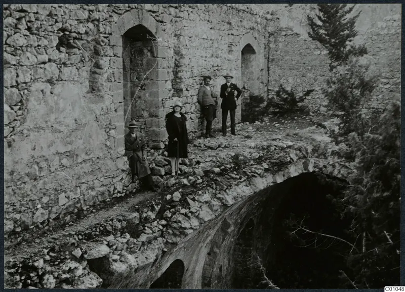 Four people standing and sitting on a rubble ledge beside a ruined stone wall with arched openings, conifer trees at right