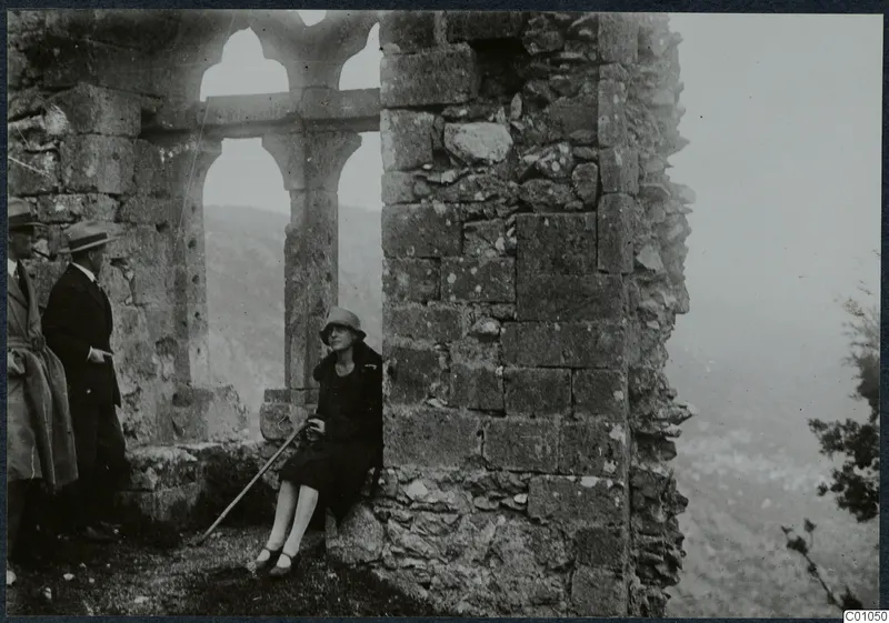 Two men stand beside a ruined stone arched window while a woman sits against a wall, with hills behind